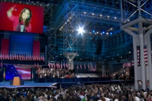 Eine große Menschenmenge sitzt vor einer Bühne mit einem Rednerpult, einem Mikrofon, Flaggen, einem Bildschirm mit dem Bild einer Frau und Scheinwerfern und Stangen während einer Präsidentschaftsdebatte.