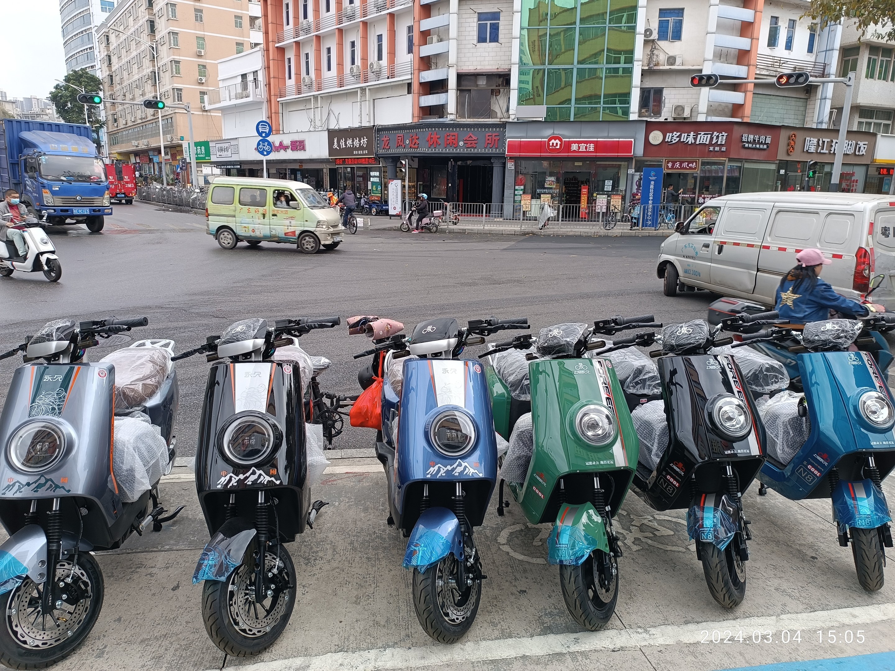 Eine Reihe von elektrischen Rollern, die auf einer Straße geparkt sind, mit einer Person in einer Mütze davor, Gebäuden, Verkehrszeichen, Bäumen und einem klaren blauen Himmel im Hintergrund.