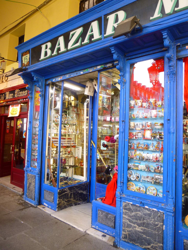 Ein Ladengeschäft mit einer blauen Tür und einem Schild, auf dem "Bazar" steht, das verschiedene Gegenstände in Schränken zeigt und so den Eindruck eines belebten Bazars erweckt.