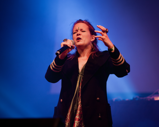 Eine Frau in einem schwarzen Kleid singt auf der Bühne ins Mikrofon, beleuchtet von blauem Licht, ihre Augen geschlossen und ihr Gesichtsausdruck voller Emotion.