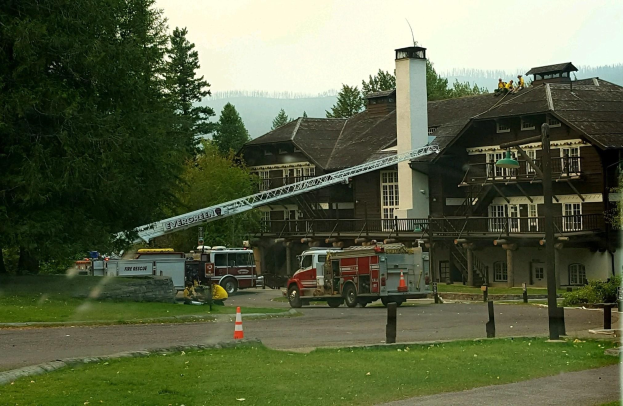 Feuerwehrfahrzeug vor einem großen Gebäude mit Fenstern, Geländern, Säulen und einem Schornstein geparkt, mit Verkehrskegeln, Gras, Bäumen, Laternen und einigen Menschen zu sehen; Hügel und ein klarer blauer Himmel im Hintergrund.