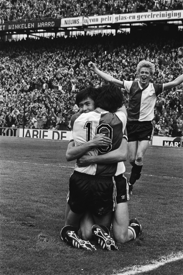 Drei Fußballspieler umarmen sich auf dem Boden und feiern ein Tor vor einer jubelnden Menge in einer schwarz-weißen Stadionszene.