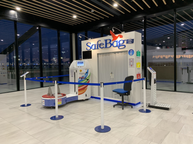 Sicherheitskontrolle für Gepäckstücke am Flughafen mit einem Stuhl, Absperrgittern, Maschinen, einem Bildschirm, einer bedruckten Tafel, Glaswänden, Deckenlampen und sichtbarem Boden.
