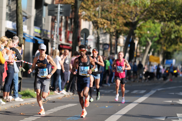 Gruppe von Läufern bei einem Marathon auf einer Stadtstraße, mit Zuschauern auf der linken Seite, Bäumen, Gebäuden und einem klaren blauen Himmel im Hintergrund; alle Läufer tragen Turnschuhe und Hüte.