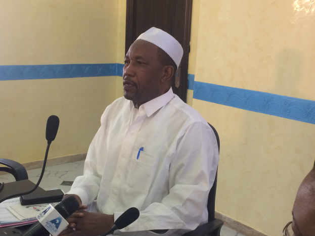 Ein Mann in einem weißen Kleid und Hut spricht an einem Tisch mit Mikrofonen, Papieren und einer anderen Person mit Brille in der Nähe in einem Raum mit einer Tür im Hintergrund.