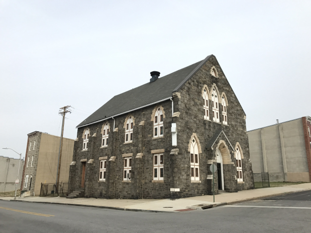 Alte steinerne Kirche an einer Straßenecke mit umliegenden Gebäuden, Straßeninfrastruktur und Fahrzeugen unter einem klaren blauen Himmel.