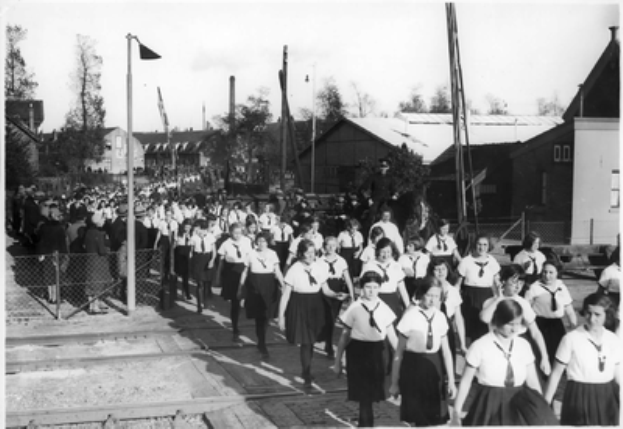 Eine Gruppe von Mädchen in Schuluniformen marschiert auf einer Straße, mit einem Zaun auf der linken Seite und Menschen dahinter, Pfählen, Bäumen und Gebäuden im Hintergrund und einem sichtbaren Himmel, in Schwarz-Weiß.