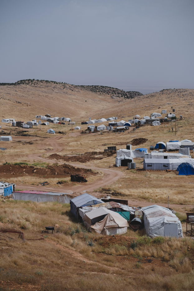 Eine Gruppe von Zelten auf einem Schmutzfeld umgeben von Gras und Pfählen, mit einem Hügel im Hintergrund und einem klaren blauen Himmel.