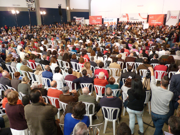 Ein volles Stadion mit vielen sitzenden und stehenden Menschen, eine B√ľhne mit zwei Rednern auf einem Podium und mehrere Banner und Lichter in der N√§he.