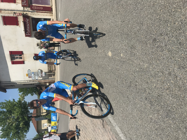 Eine Gruppe von Radfahrern fährt eine Straße entlang an einem Gebäude mit Fenstern und Tafeln mit Text vorbei, mit Bäumen und einem klaren Himmel im Hintergrund und Zuschauern, die auf der Straße darunter stehen.