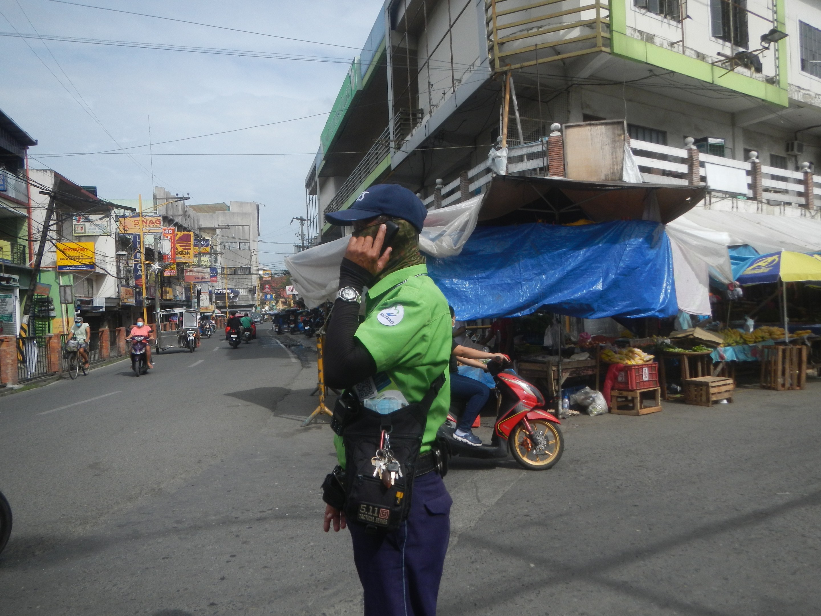 Ein Polizist in grüner und blauer Uniform und Mütze steht auf einer Straße und spricht in ein Handy, mit Fahrzeugen, Marktständen und einem Gebäude im Hintergrund unter einem bewölkten Himmel.