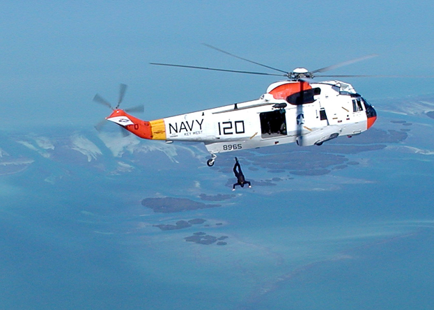Retterhubschrauber fliegt über Wasser mit einer Person darunter, Himmel im Hintergrund.