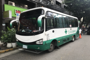 Ein grüner und weißer Doppeldeckerbus auf einem Parkplatz neben einem Gebäude geparkt, mit Fahrzeugen auf der linken Seite und Pflanzen und einer wandmontierten Tafel auf der rechten Seite.