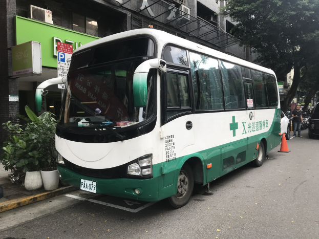 Ein grüner und weißer Doppeldeckerbus auf einem Parkplatz neben einem Gebäude geparkt, mit Fahrzeugen auf der linken Seite und Pflanzen und einer wandmontierten Tafel auf der rechten Seite.