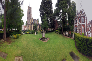 Ein Park mit einem zentralen Brunnen, umgeben von Rasen, Pflanzen, Bäumen und Gebäuden mit Fenstern, mit einem prominenten Baum in der Mitte, Fahrzeugen auf der angrenzenden Straße und einem klaren blauen Himmel im Hintergrund.
