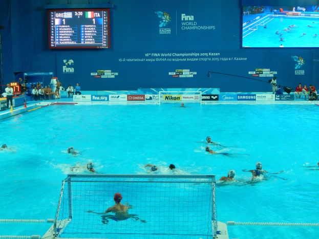 Eine Gruppe von Menschen spielt Wasserball in einem großen Pool mit einem Netz in der Mitte, Zuschauern auf der linken Seite und einer Wand im Hintergrund mit Text und Logos für die 2016 FINA World Championships in Kazan.