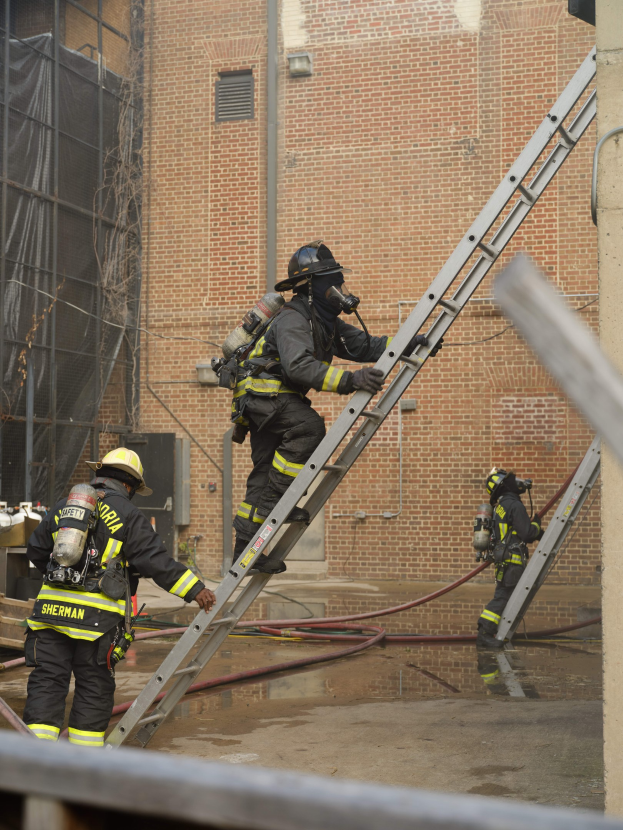Feuerwehrleute in Helmen und Ausrüstung klettern an einer Leiter vor einem Backsteingebäude mit sichtbaren Rohren und einem Metallstab an der Basis hoch, während im Hintergrund ein weiteres Gebäude mit Fenstern und einem Netz zu sehen ist.