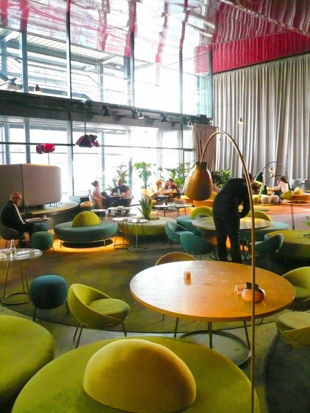 Ein geräumiger Hotellounge mit grünen Stühlen, Tischen, Sofas, stehenden Menschen, Pflanzen, einer Stehlampe, Vorhängen und Deckenleuchten.