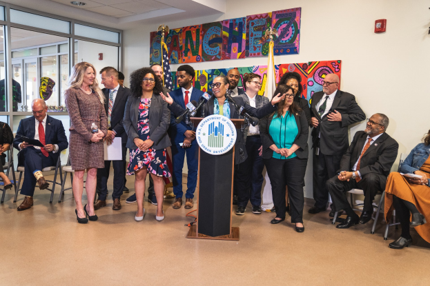 Gruppe von Menschen vor einem Rednerpult mit Mikrofon und einer Tafel mit dem Text 'Sancho City Councilman Joins City Council', mit sitzenden und stehenden Teilnehmern, Flaggen, Feuerlöscher und Deckenleuchten im Hintergrund.