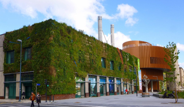 Großes nachhaltiges Gebäude mit einer grünen Wand, umgeben von städtischer Infrastruktur einschließlich Straßenlaternen, Verkehrssignalen, Fahrzeugen, Fußgängern, Bäumen und einem bewölkten Himmel.