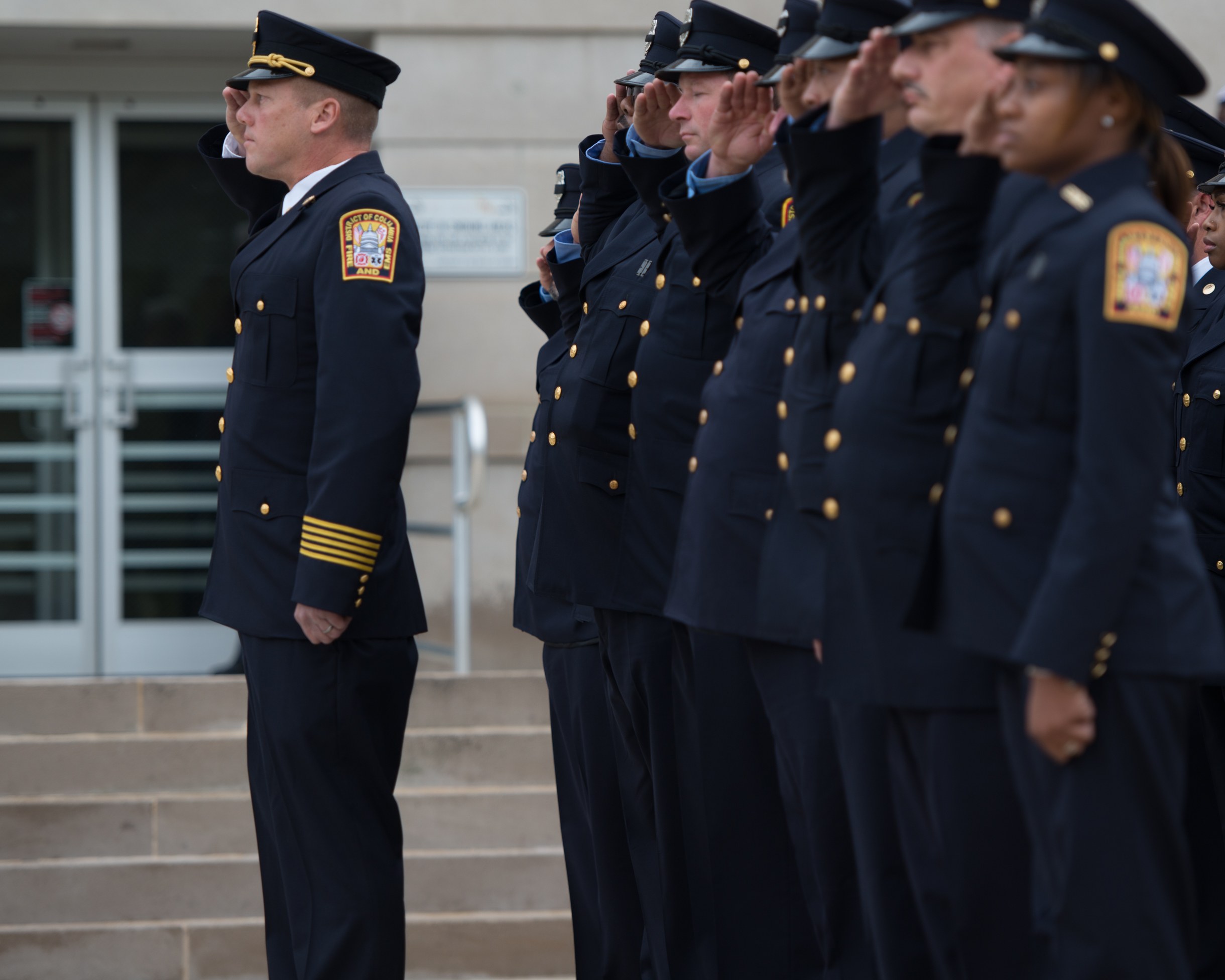 Gruppe von Polizisten in Uniform und Mützen, die in Formation stehen und vor einem Gebäude mit Glastüren und Treppen salutieren.