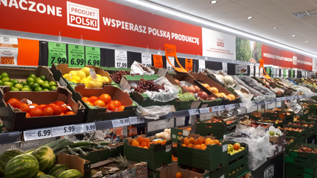 Ladeninneres mit Kartonboxen verschiedener Obst- und Gemüsesorten, jede mit Preisschildern, Text- und Bildtafeln an den Wänden und Deckenbeleuchtung.