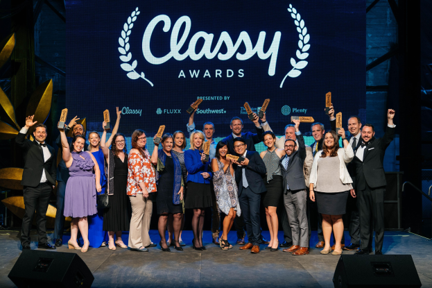 Gruppe von Menschen auf einer Bühne mit Auszeichnungen, mit Lautsprechern darunter und einem Bildschirm im Hintergrund mit der Aufschrift "Classy Awards 2019"