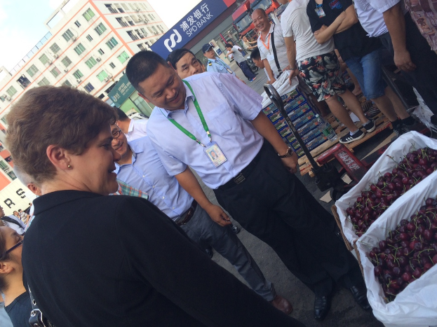 Eine Straßenszene mit Menschen, die auf der Straße stehen, einem Korb mit Kirschen unten rechts, Kisten dahinter, Gebäuden im Hintergrund mit Texttafeln und dem Himmel oben links.
