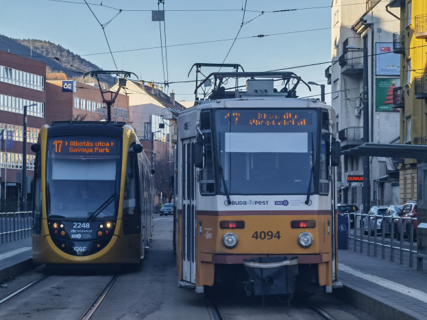 Zwei gelbe und weiße Straßenbahnen fahren auf Stadtbahngleisen neben hohen Gebäuden, mit Fahrzeugen, Geländern, Bäumen und Gebäuden im Hintergrund unter einem Himmel.