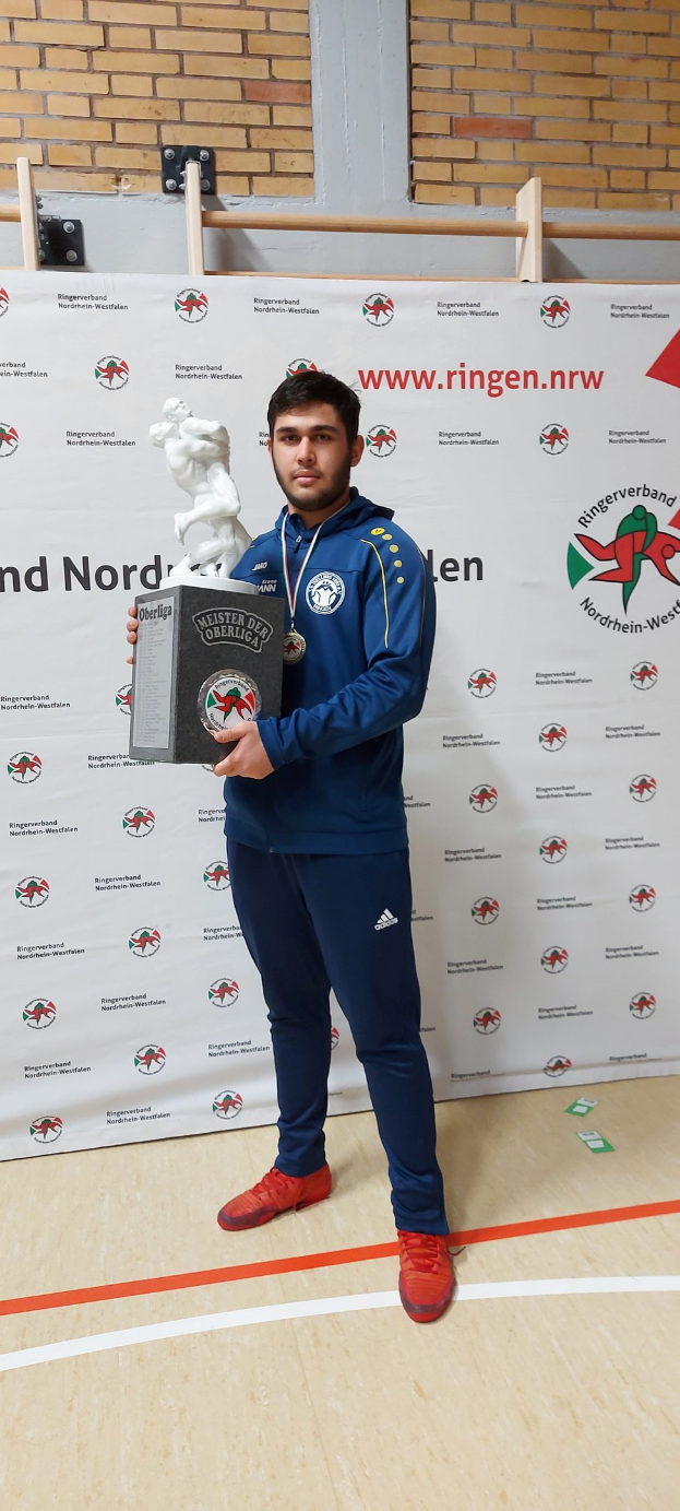 Ein Mann in einem Anzug hält einen Pokal und trägt eine Medaille, vor einer geschmückten Wand stehend.