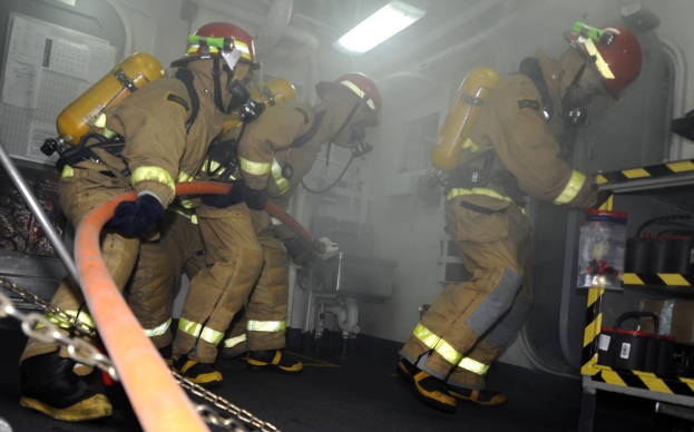 Feuerwehrleute in Schutzausrüstung und Helmen arbeiten an einem Feuerwehrauto, mit Equipment-Regalen und Hintergrundinfrastruktur im Hintergrund.