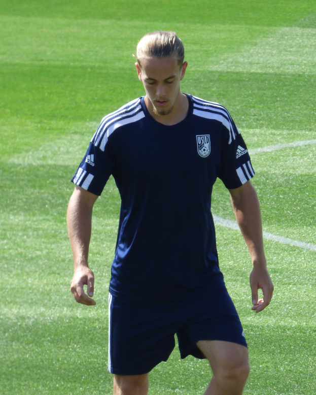 Ein Mann in einem blauen T-Shirt und Shorts schreitet selbstbewusst über ein grünes Feld und sieht aus wie ein professioneller Füßballspieler mit entschlossener Miene.