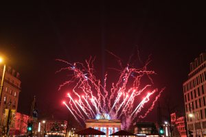 Eine belebte Stadtstraße in Berlin an Silvester, voller Menschen, Fahrzeuge und Gebäude, beleuchtet von Lichtern und Feuerwerk in einer festlichen Atmosphäre.