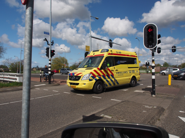 Ein Rettungswagen fährt auf einer Straße neben einer Ampel, mit einem Radfahrer im Vordergrund und Gebäuden, Bäumen und Lichtmasten im Hintergrund unter einem bewölktem Himmel; ein Spiegel ist unten sichtbar.
