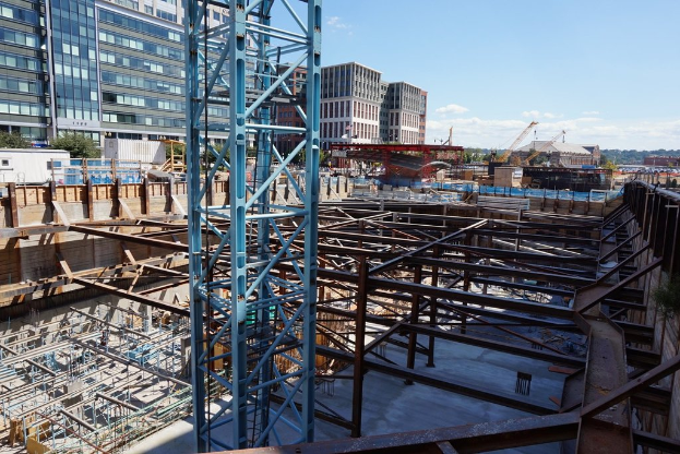 Das Bild zeigt eine Baustelle mit Gebäuden und Büros links, Eisenstangen und Maschinen rechts und verschiedene Geräte und Kräne in der Mitte, unter einem Himmel oben rechts.