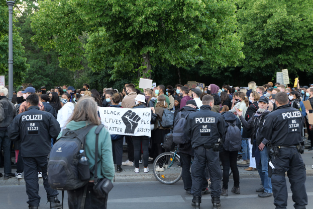 Eine große Gruppe von Menschen steht an der Straße, einige halten Schilder, mit einem Fahrrad im Vordergrund und Bäumen und einem Pfahl im Hintergrund, bei einer Black-Lives-Matter-Demonstration in Berlin.