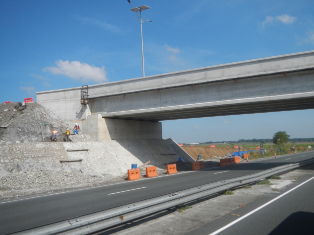 Autobahn mit Brücke darüber, ein darunter fahrendes Auto und Bauarbeiter am Straßenrand, umgeben von Verkehrskegeln, Steinen und einem Lichtmast, mit Bäumen und einem bewölkten Himmel im Hintergrund.