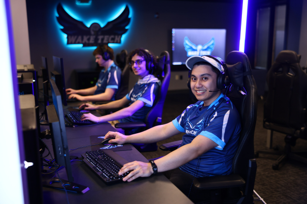 Gruppe von Menschen an Computern mit Headsets, die während eines Esport-Turnieraufbaus mit Monitoren, Tastaturen und einem Logo an der Wand lächeln.