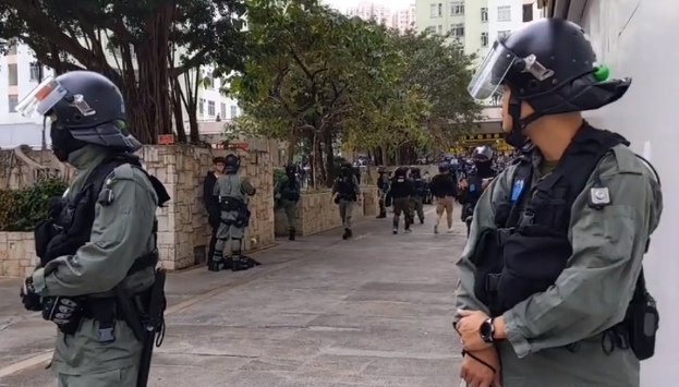 Polizeibeamte in Helmen und Schutzwesten stehen in einer Reihe auf einer Straße in Hong Kong, mit Bäumen, Gebäuden und Himmel im Hintergrund.
