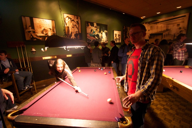 In einem Club spielen zwei Personen Snooker an einem von zwei Tischen, während mehrere andere im Hintergrund stehen und sich unterhalten, vor einer cremefarbenen Wand mit Foto-Postern.