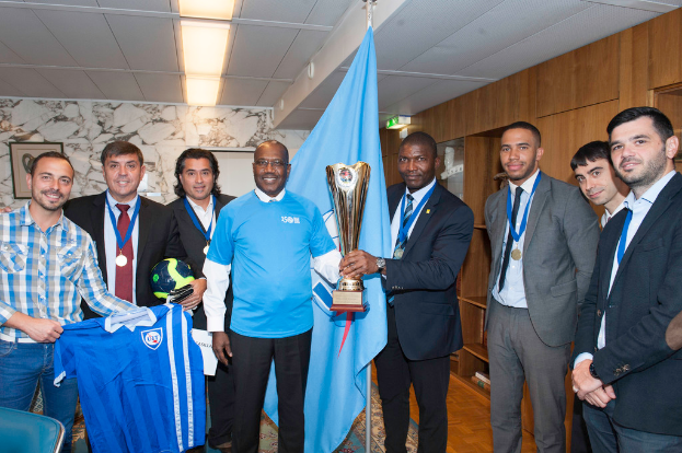 Eine Gruppe von Menschen, die zusammenstehen, wobei zwei in der Mitte einen Pokal halten, einer links ein T-Shirt und ein weiterer links einen Ball, und eine Flagge im Hintergrund.