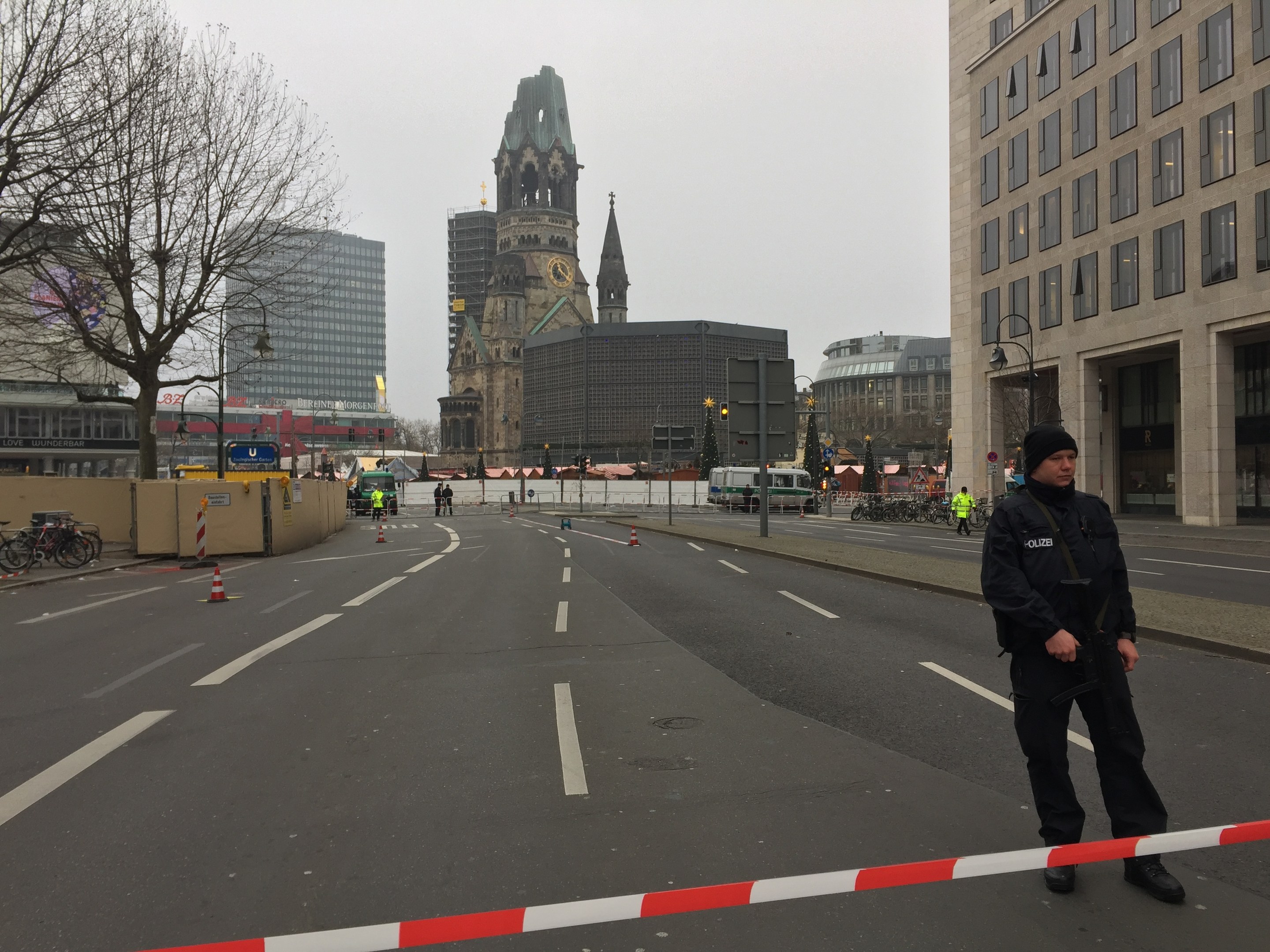 Ein Polizist in schwarzer Uniform und Mütze steht in einer Berliner Straße umgeben von Verkehrskegeln, Fahrrädern, Laternenpfählen, Bäumen und Gebäuden, mit einem Uhrenturm und blauem Himmel im Hintergrund.