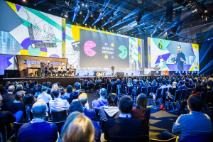 Eine große Menschenmenge, die auf Stühlen vor einer Bühne sitzt, mit einigen Individuen, die auf dem Podium in der vorderen Reihe stehen, einem Tisch mit verschiedenen Gegenständen auf der linken Seite und Bildschirmen und Lichtern im Hintergrund auf der Deutschen Internet-Konferenz 2019.