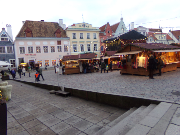 Ein belebter Weihnachtsmarkt in Tallinn, Estland mit Menschen um geschmückte Stände, festlich geschmückte Gebäude im Hintergrund, bewölktem Himmel, Treppe nach oben und Topfpflanzen.