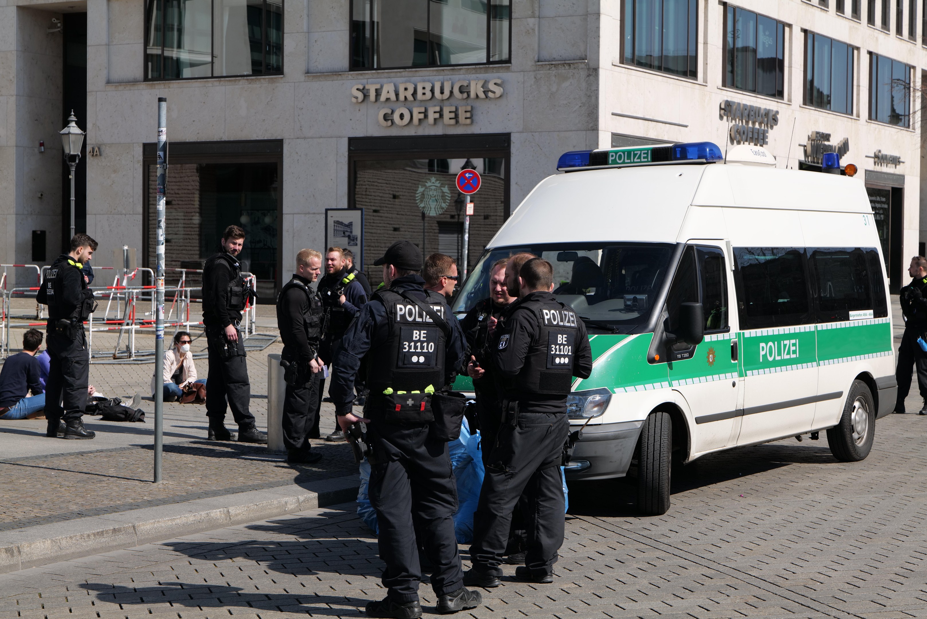 Eine Gruppe von Polizisten steht vor einem Starbucks-Kaffeehaus, rechts ein Van und links ein paar Menschen; im Hintergrund sind ein Gebäude, ein Schild, ein Lichtmast und ein Zaun zu sehen.