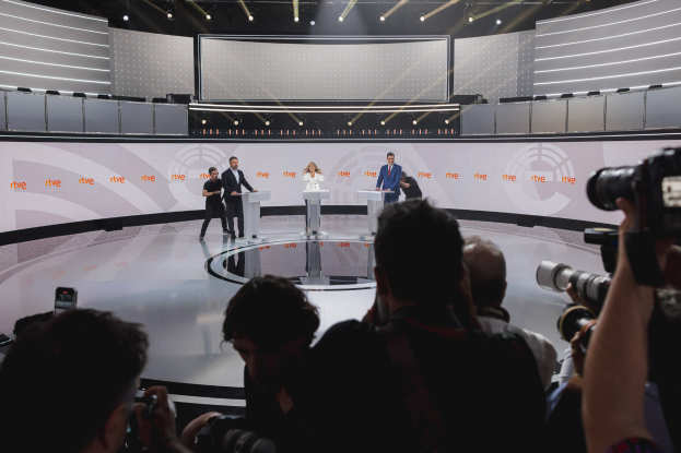 Eine Gruppe von Menschen steht auf einer Bühne mit zwei Podesten, die in eine Diskussion vertieft sind, während andere Kameras auf beiden Seiten halten und eine beleuchtete Wand im Hintergrund zu sehen ist.