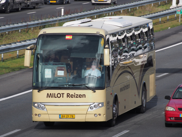 Ein Minibus mit Passagieren auf einer Autobahn neben einem roten Auto, mit Geländern und Gras am Straßenrand und Texttafeln im Hintergrund.