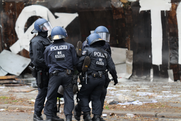 Polizeibeamte in schwarzen Uniformen und blauen Helmen stehen vor einem Graffiti-bedeckten Gebäude während einer Krawall, mit verstreuten Papieren und Gegenständen auf dem Boden.