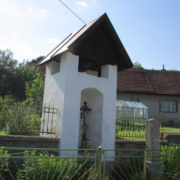 Kleine weiße Kirche mit einem Kreuz darauf, umgeben von einem Metallzaun, Pflanzen, Gras, einem Weg, nahen Häusern, Polen, Drähten und einem bewölkten Himmel, identifiziert als die Kirche des Heiligen Grabes.