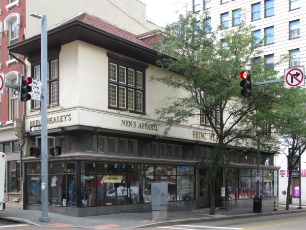 Ecke einer belebten Stadtstraße mit einem geparkten Bus, Gebäuden, Bäumen, Laternen, Verkehrsampeln, Schildern und einem Kaufhaus mit Schaufensterpuppen in Kleidern, mit dem Himmel im Hintergrund.